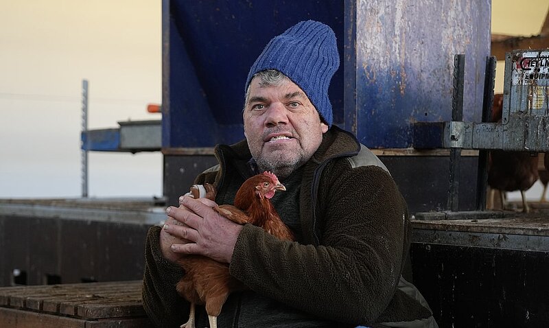 Ein Mann mit einem blauen Mütze sitzt auf einem Holzstapel und hält ein Huhn in seinen Armen. Er blickt direkt in die Kamera und hat einen ernsten Ausdruck im Gesicht. Im Hintergrund sind blasse Lichter und eine Struktur, die auf einen Stall hinweist.