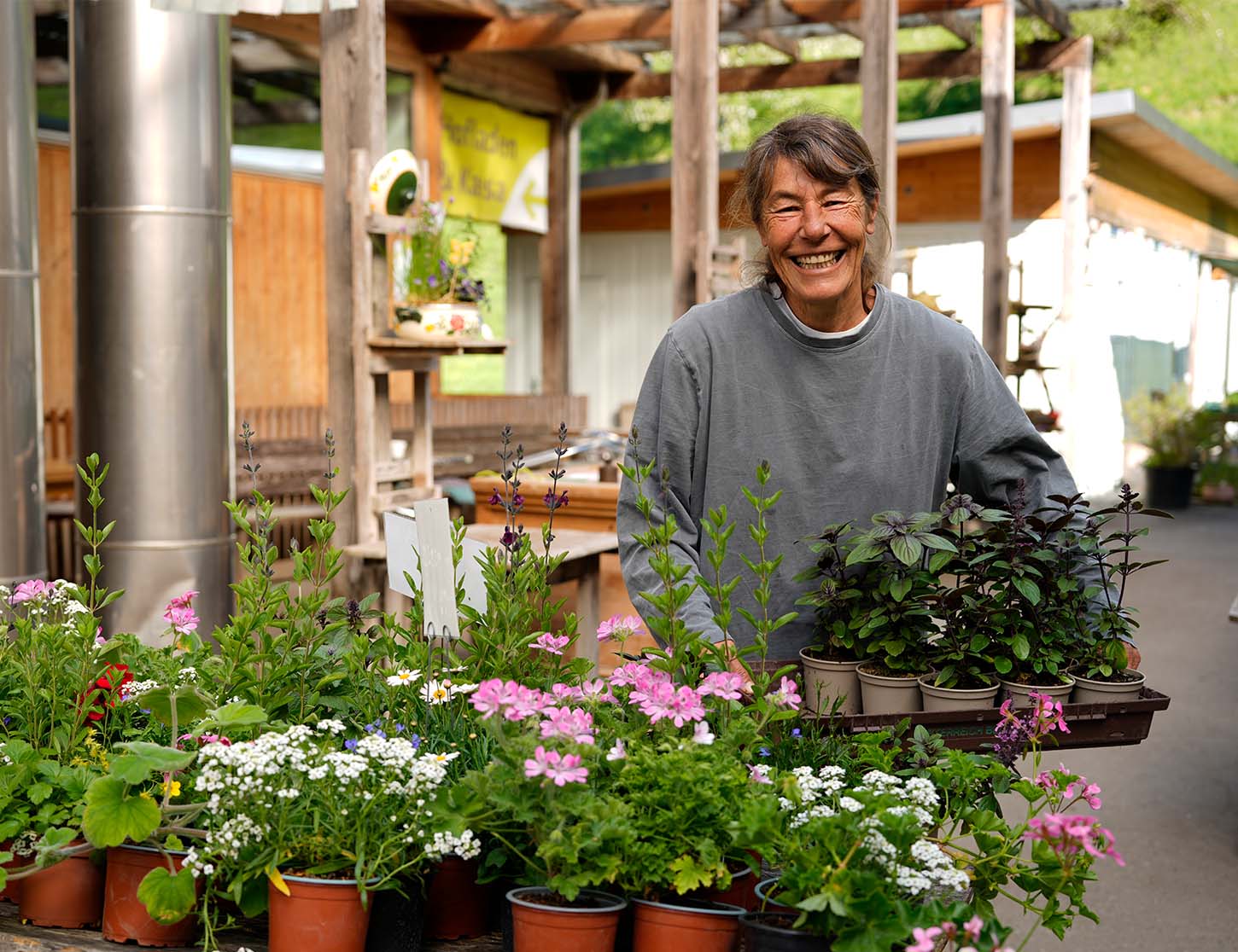 Eine lächelnde Frau steht in einem Gartenmarkt und hält eine Trage mit Pflanzen und Blüten in Töpfen. Um sie herum wachsen verschiedene Blumen, darunter Rosa- und weiße Blüten. Im Hintergrund sind Holzkonstruktionen und Pflanzen zu sehen.