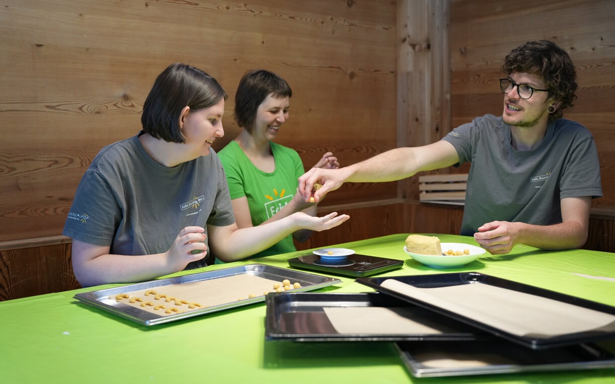 Drei Personen sitzen an einem Tisch mit grünem Tischdecken und arbeiten gemeinsam an einem Backprojekt. Eine Person reicht einer anderen eine kleine Kugel, während die dritte Person lächelt. Auf dem Tisch sind Blechtafeln mit ungleichmäßigen kleinen Keksen.