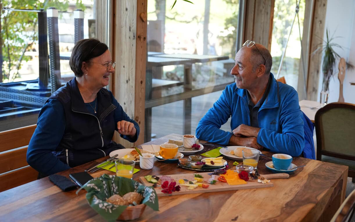 Zwei Personen sitzen an einem Tisch, lächeln und unterhalten sich. Auf dem Tisch sind verschiedene Frühstücksgerichte angerichtet, darunter Brote, Käse und Getränke. Der Raum hat große Fenster, durch die Tageslicht strömt, und die Atmosphäre ist freundlich und einladend.