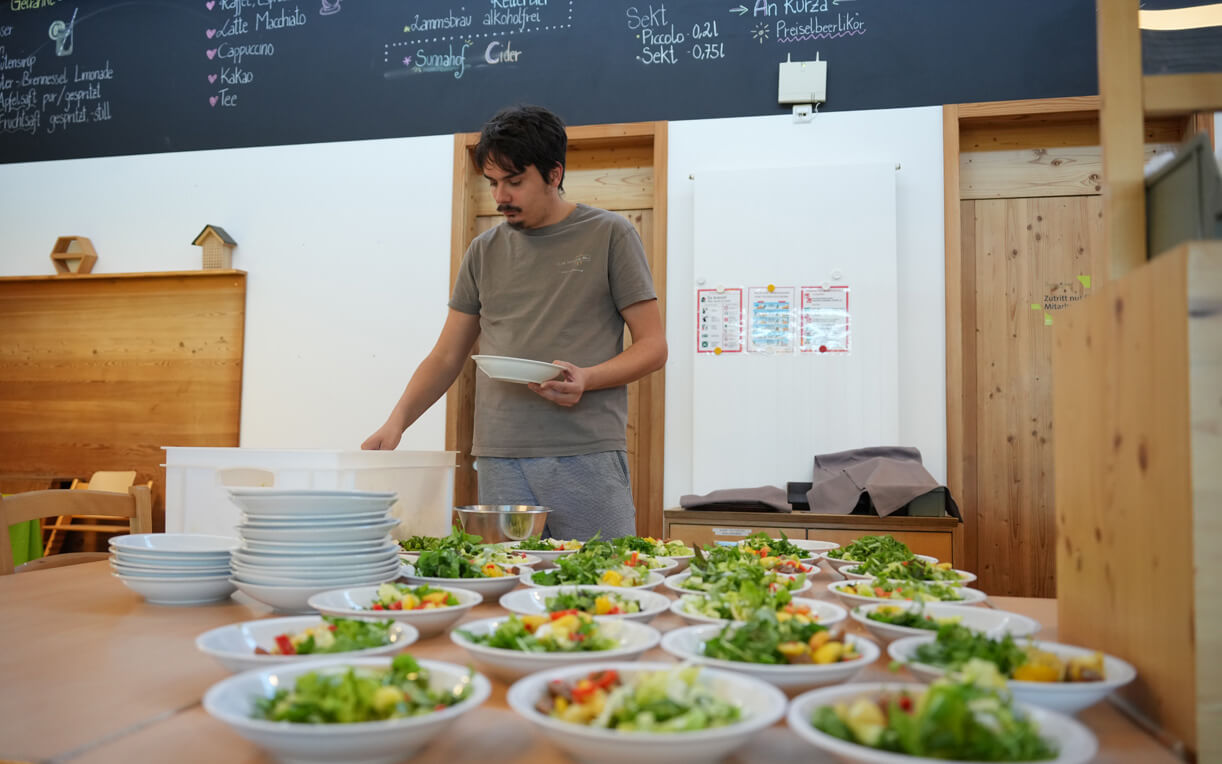 Ein junger Mann steht an einem Tisch und bereitet Schüsseln mit frischem Salat vor. Vor ihm sind mehrere weiße Teller mit bunten, frischen Zutaten angerichtet. Im Hintergrund sind Wandtafeln mit Getränken und Informationen sichtbar.
