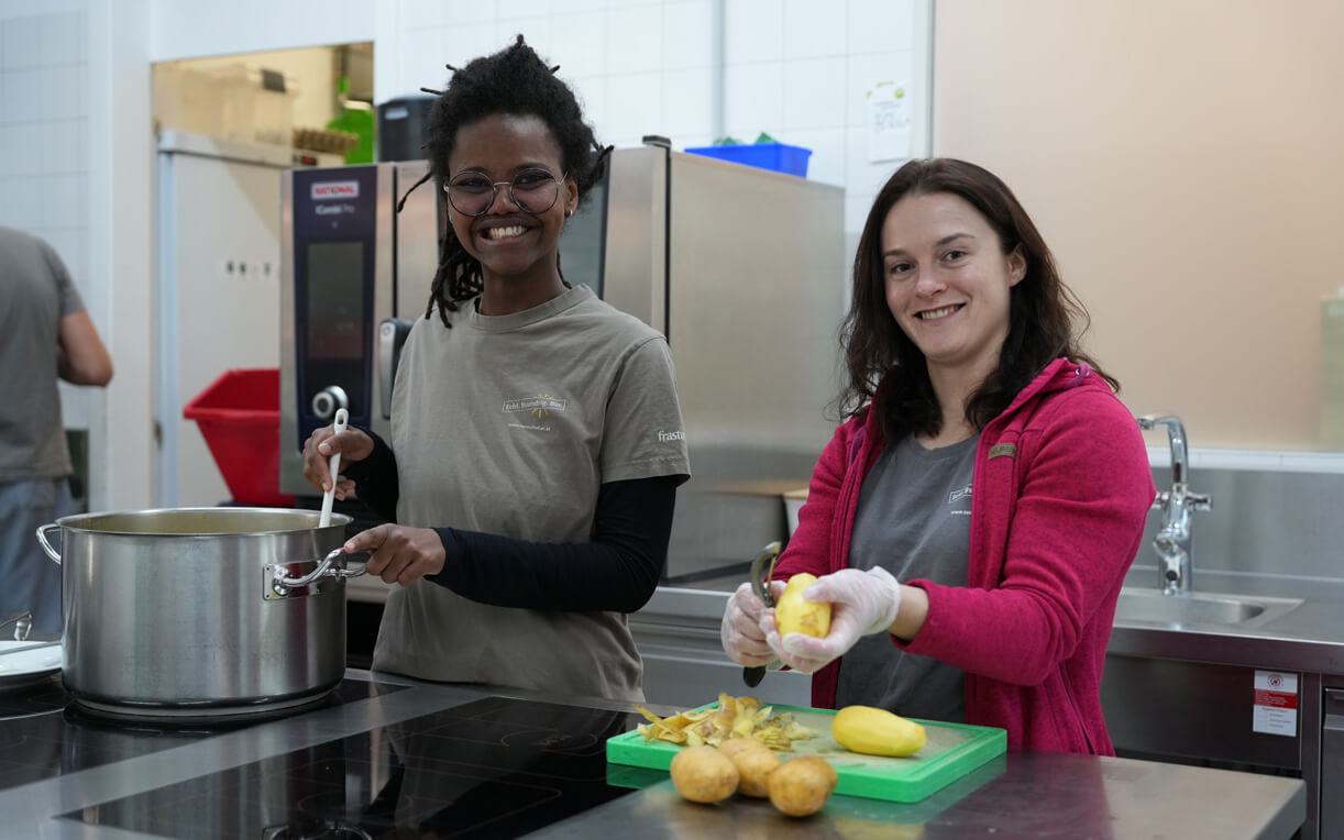 Zwei Frauen stehen in einer Küche. Eine rührt in einem großen Topf, während die andere mit Handschuhen Kartoffeln schält. Beide lächeln und sind in entspannter Arbeitsatmosphäre beschäftigt. Im Hintergrund sind Küchenelemente sichtbar.