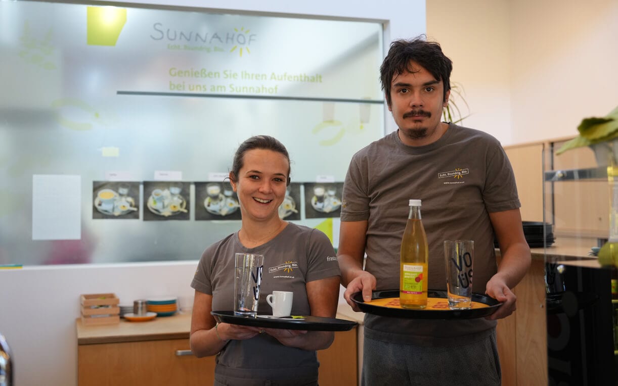 Zwei Servicemitarbeiter stehen in einem Café. Die Frau hält ein Tablett mit einem Glas und einer Tasse, während der Mann ein weiteres Tablett mit einer Flasche Getränk zeigt. Im Hintergrund ist eine Wand mit einem Logo und Informationen über den Aufenthalt im Sonnenhof zu sehen.