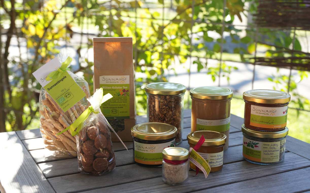 Das Bild zeigt eine Auswahl an verschiedenen Lebensmitteln auf einem Tisch. Es sind Gläser mit Honig, Kräutern und einer Verpackung für Kräutertee sowie Tüten mit Keksen und Snackstäbchen zu sehen. Der Hintergrund ist grün und sonnig, was eine entspannte Atmosphäre schafft.