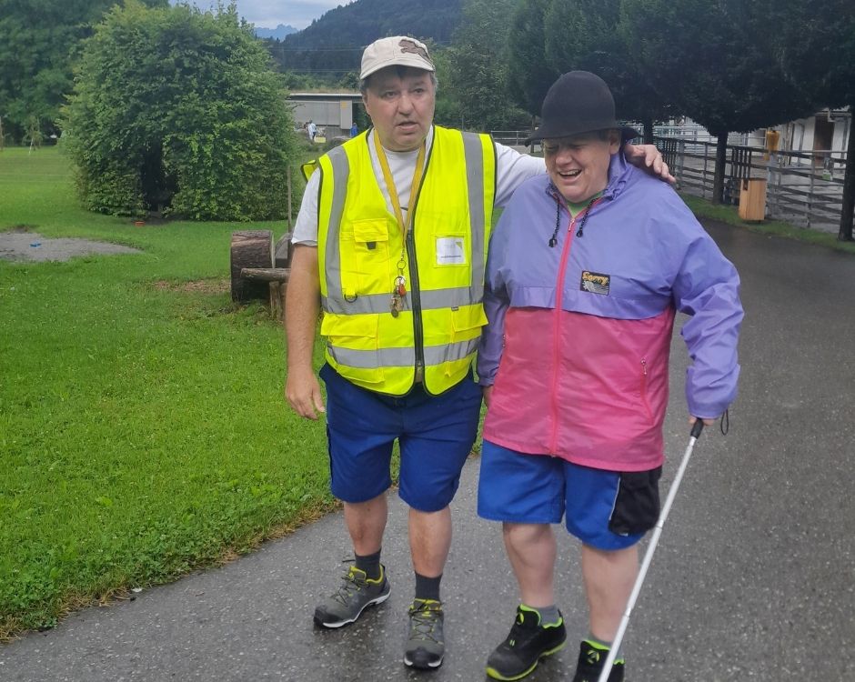 Eine Person in einer gelben Warnweste begleitet eine andere, die einen Stock benutzt, auf einem Weg in einem grünen, offenen Bereich. Die begleitete Person trägt eine bunte Jacke und lächelt, während sie zusammen gehen.