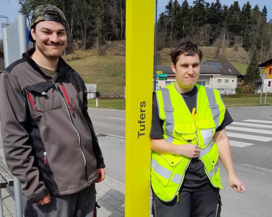Zwei junge Männer stehen an einer Haltestelle. Der links trägt eine graue Jacke und eine Mütze, der rechts eine leuchtend gelbe Warnweste. Hinter ihnen ist eine Straße mit einer Kreuzung sowie Bäume und Häuser im Hintergrund sichtbar. An einem Pfosten steht der Ort "Tufers".