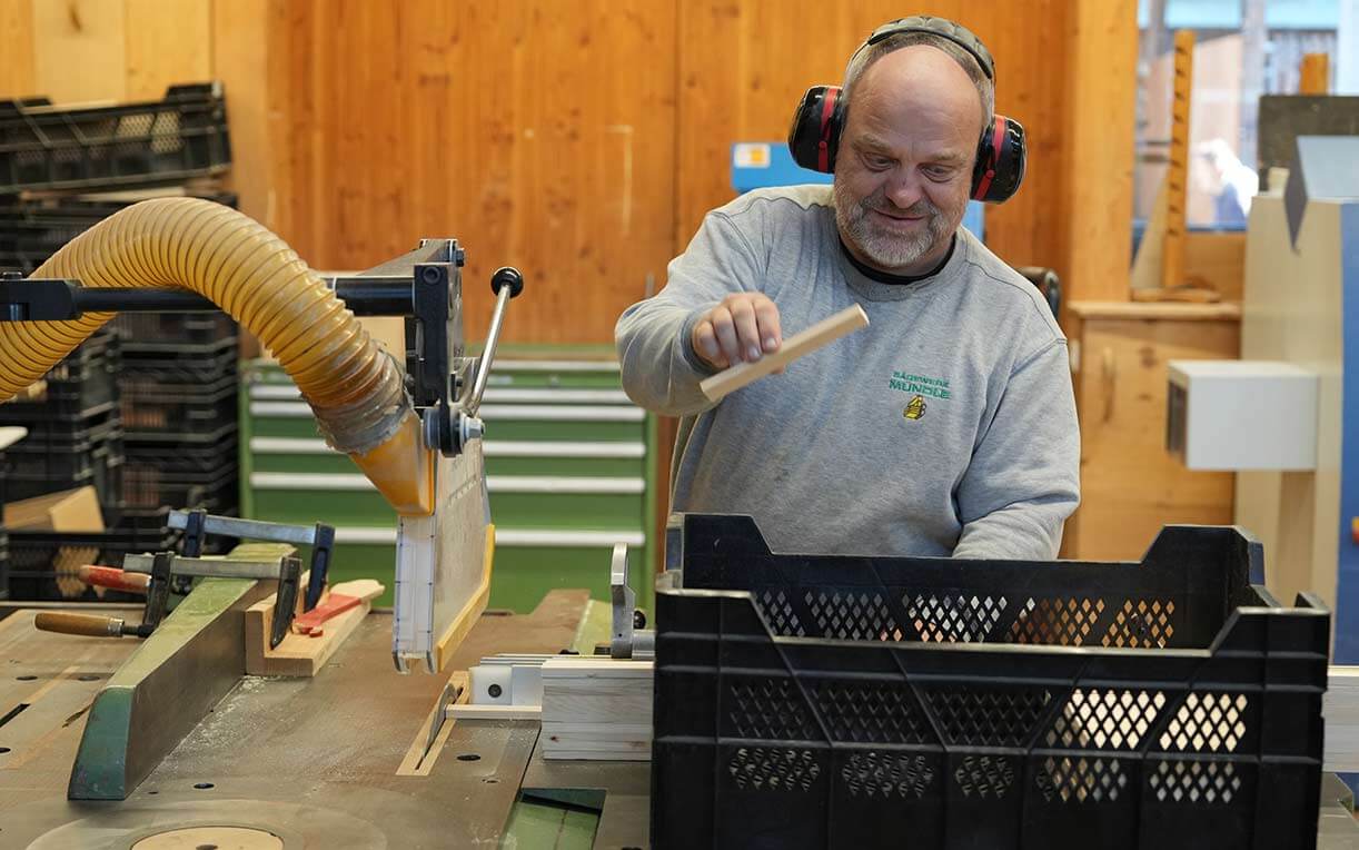 Ein Handwerker arbeitet in einer Werkstatt mit einer Tischsäge. Er trägt Gehörschutz und schneidet ein Holzstück. Im Hintergrund sind weitere Arbeitsutensilien und Holz gestapelt. Die Atmosphäre ist industriell und gut beleuchtet.