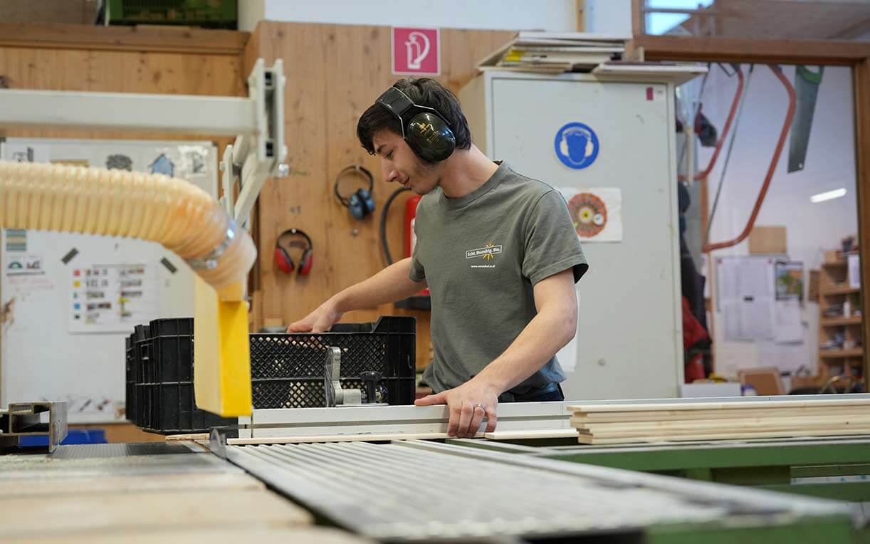 Ein junger Mann mit Ohrschutz arbeitet in einer Holzwerkstatt. Er bedient eine Maschine zum Schneiden von Holz und konzentriert sich auf seine Aufgabe. Im Hintergrund sind Werkzeuge und Materialien sichtbar, die zur Holzbearbeitung verwendet werden.