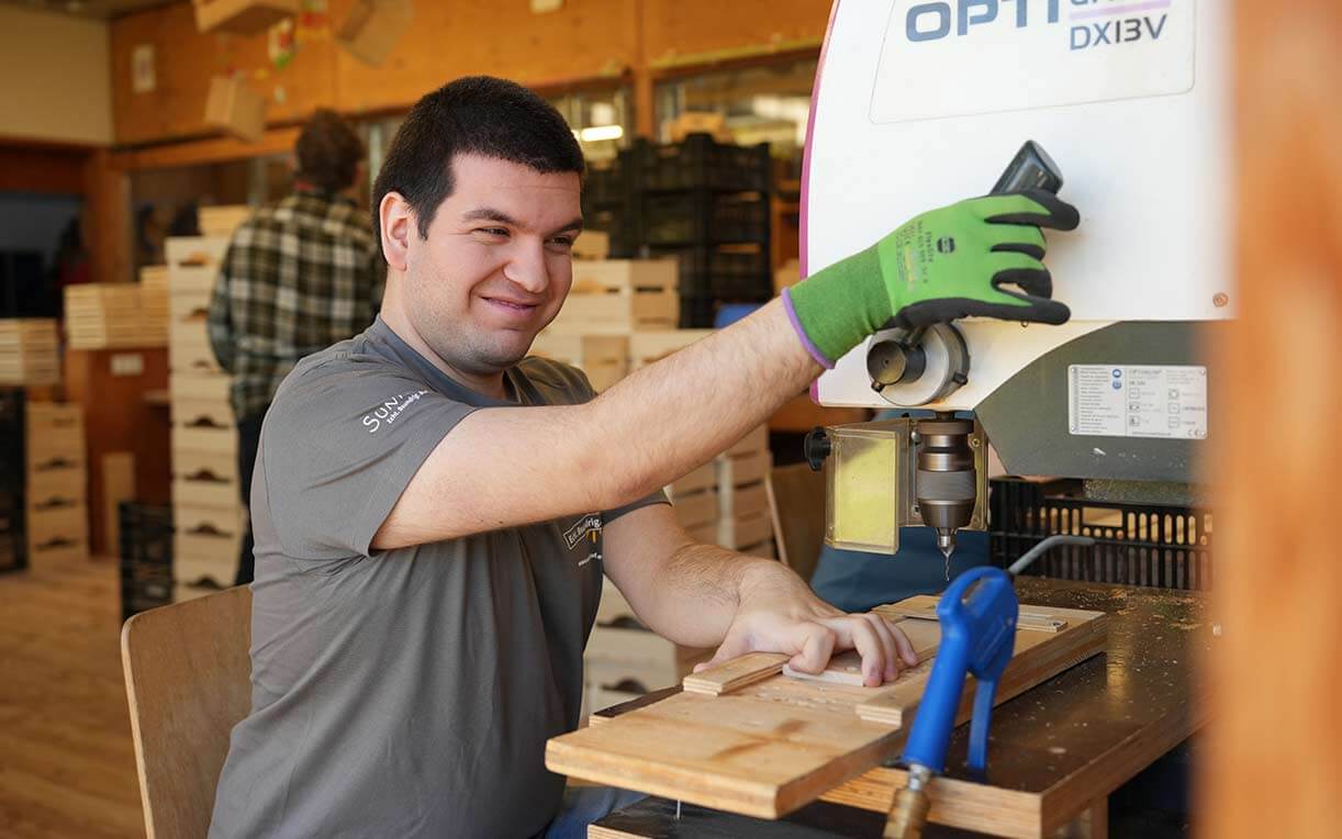 Ein junger Mann sitzt an einer Werkbank und bedient eine Holzfräse. Er lächelt, während er mit einer Hand das Werkstück hält und mit der anderen die Maschine steuert. Im Hintergrund sind Holzboxen und weitere Personen zu sehen.