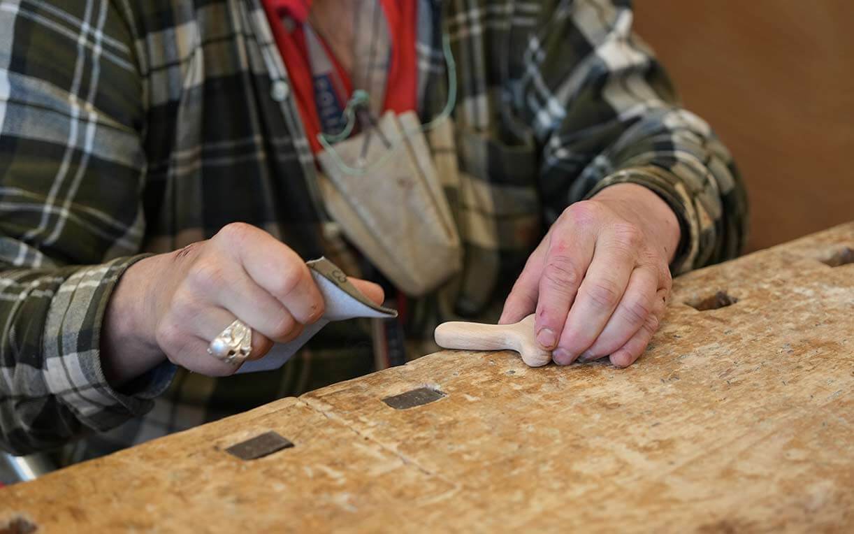 Ein Handwerker arbeitet mit einem kleinen Holzteilstück auf einer Holzoberfläche. Mit einem Werkzeug in der linken Hand wird die Holzfigur bearbeitet, während die rechte Hand das Stück stabil hält. Der Handwerker trägt ein kariertes Hemd und hat einen Ringe an der linken Hand.