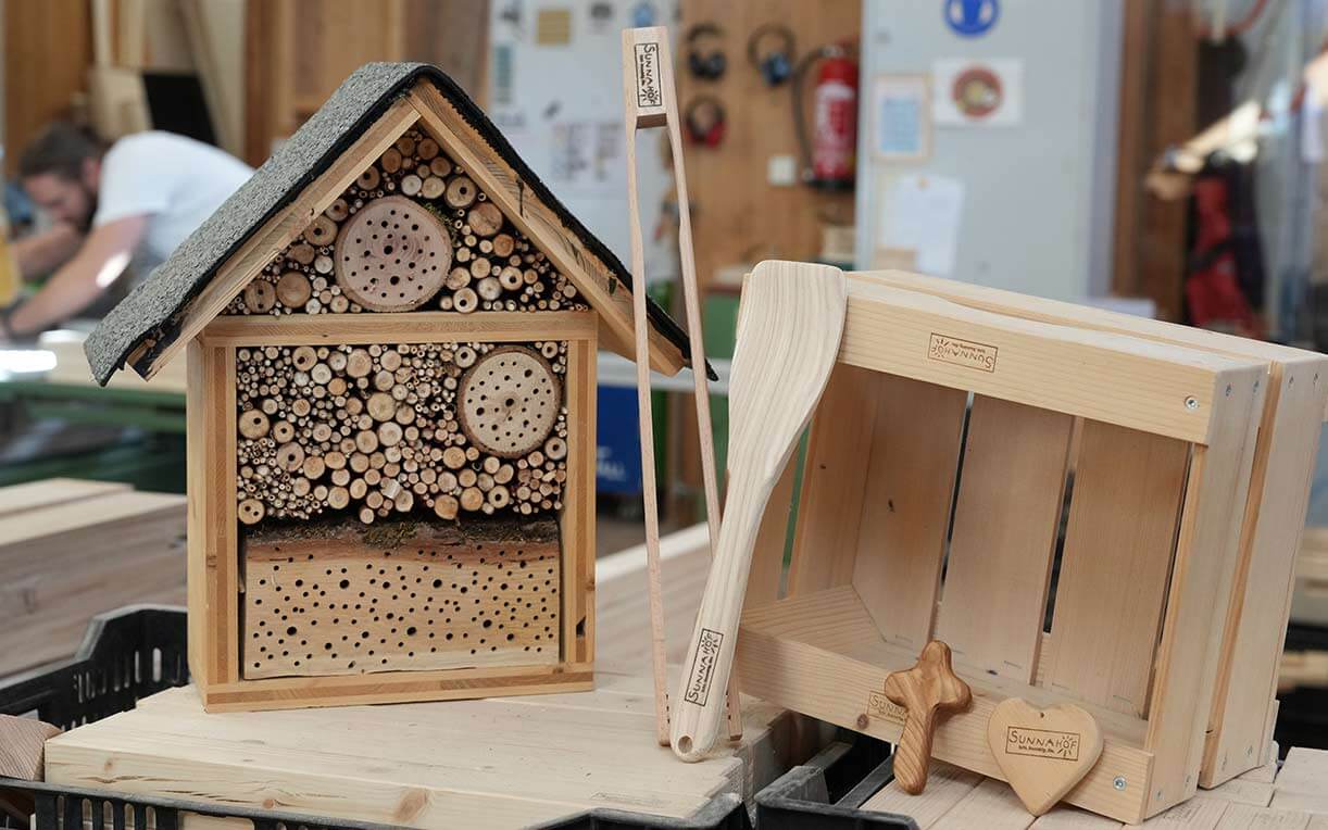 Ein Insektenhotel in Form eines kleinen Hauses, gefertigt aus verschiedenen Holzstücken mit Bohrlöchern. Daneben liegen ein hölzerner Löffel und zwei handgefertigte Holzformen in Herz- und Kreuzform. Der Hintergrund ist unscharf und zeigt eine Werkstatt.