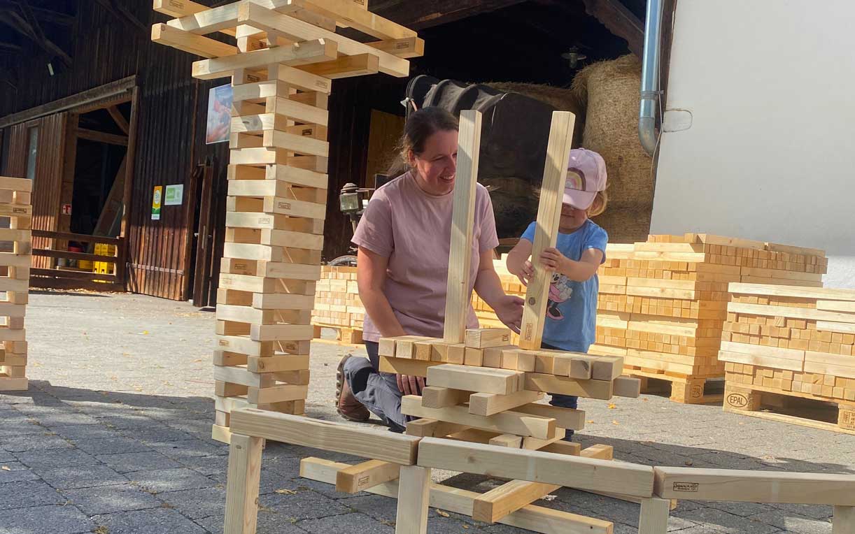 Eine Frau und ein kleines Kind bauen aus Holzklötzen eine Struktur. Die Frau sitzt auf dem Boden und hilft dem Kind, einen Block auf die Spitze der Konstruktion zu setzen. Im Hintergrund sind weitere Stapel von Holzklötzen sichtbar. Es ist ein sonniger Tag.