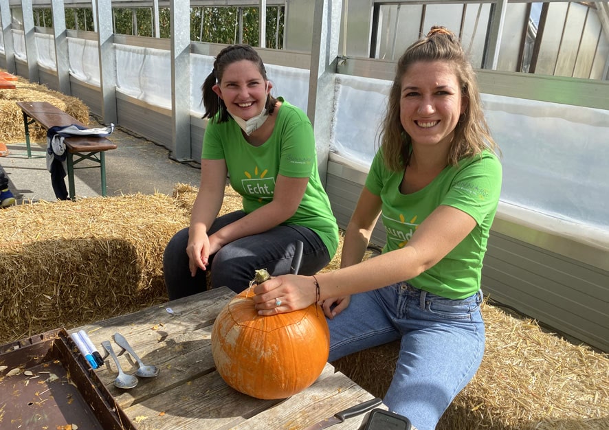 Zwei Frauen in grünen T-Shirts sitzen an einem Holztisch in einem Gewächshaus. Vor ihnen liegt ein orangefarbener Kürbis, den sie bearbeiten. Die Frauen lächeln und scheinen Spaß zu haben. Im Hintergrund sind Strohballen und eine klare, helle Umgebung sichtbar.