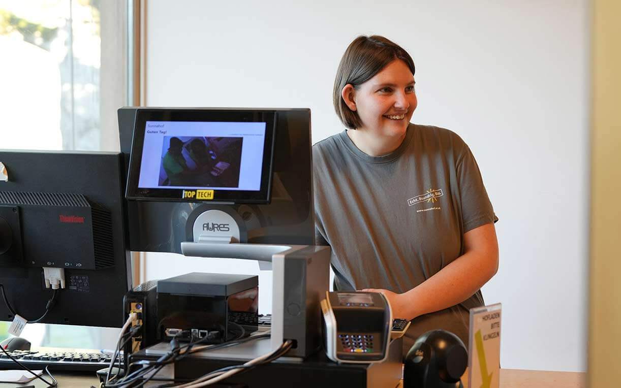 Eine lächelnde Person steht an einem Empfangstresen. Im Hintergrund sind Computerbildschirme sichtbar. Der Schreibtisch ist mit Bürogeräten ausgestattet. Die Person trägt ein graues T-Shirt und wirkt freundlich, während sie mit einem Besucher spricht.