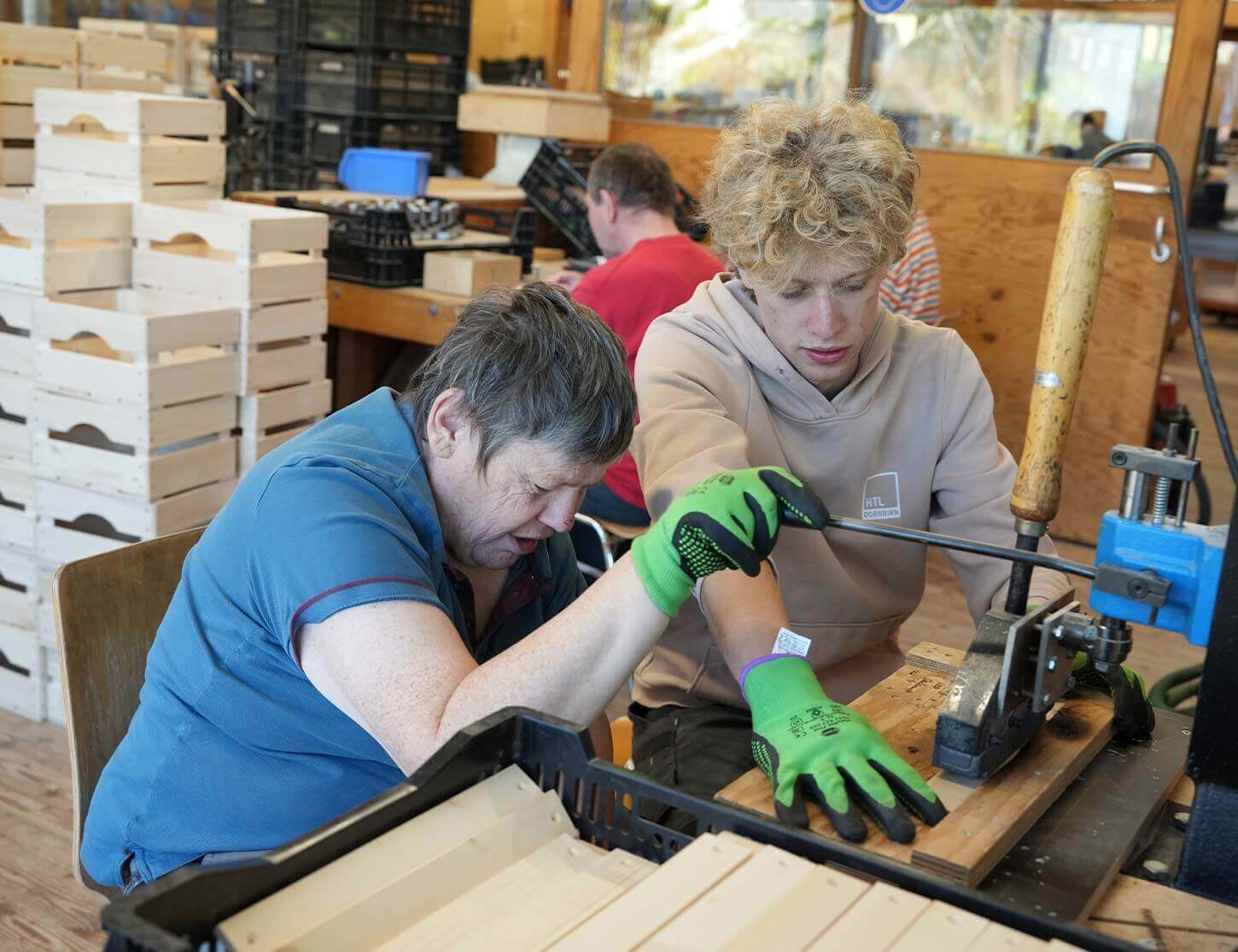 Zwei Personen arbeiten gemeinsam in einer Werkstatt. Eine Person mit kurzer Frisur und blauer Kleidung zieht mit beiden Händen an einem Werkzeug, während die zweite Person, die grüne Handschuhe trägt, aufmerksam zusieht und unterstützt. Im Hintergrund sind Holzboxen gestapelt.
