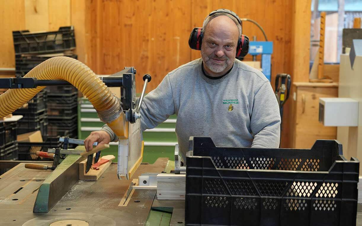Ein Handwerker arbeitet an einer Tischsäge in einer Werkstatt. Er trägt Ohrenschützer und schaut lächelnd in die Kamera, während er Holz bearbeitet. Im Hintergrund sind Holzregale und Werkzeuge zu sehen.