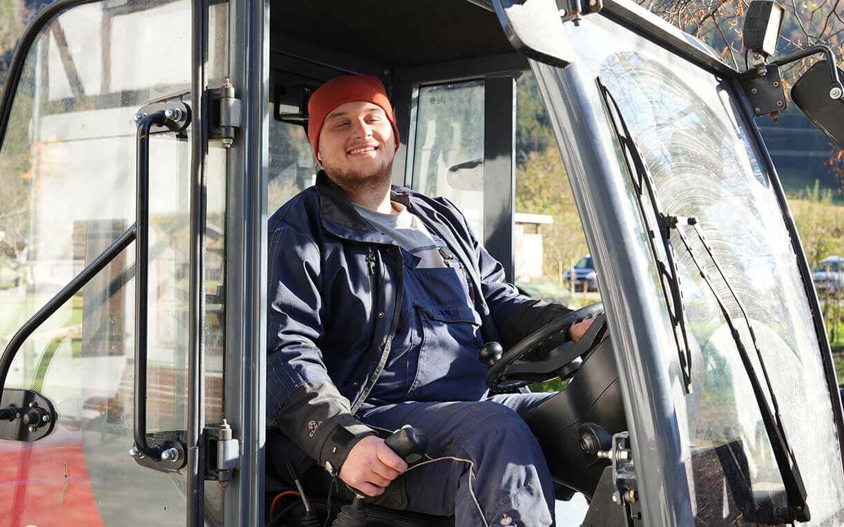 Ein lächelnder Mann sitzt in einem Traktor. Er trägt eine blaue Arbeitsjacke und eine rote Mütze. Der Traktor ist im Freien und ist teilweise von Bäumen umgeben. Das Bild vermittelt eine freundliche Atmosphäre und zeigt die Tätigkeit in der Landwirtschaft.