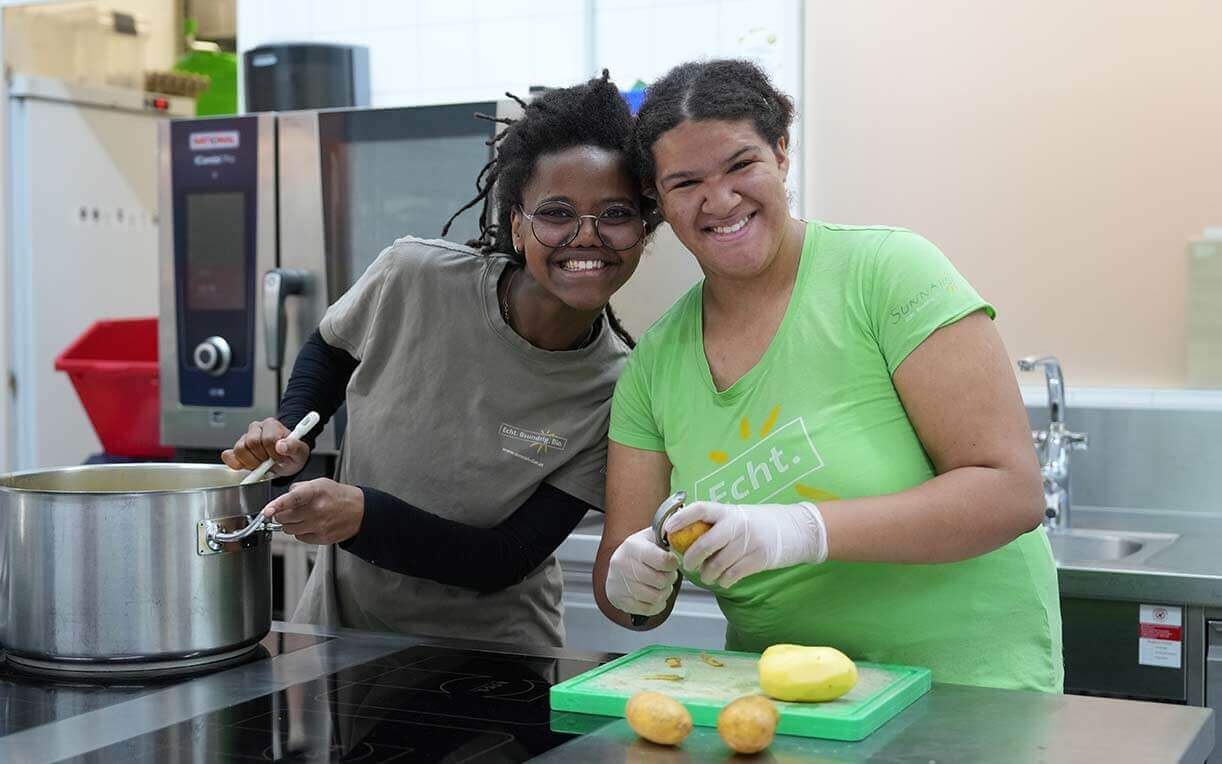 Zwei Frauen lächeln in einer modernen Küche. Eine steht neben einem großen Topf und hält einen Löffel, die andere schält eine Kartoffel auf einem Schneidebrett. Beide tragen Handschuhe und wirken zufrieden bei ihrer Arbeit.