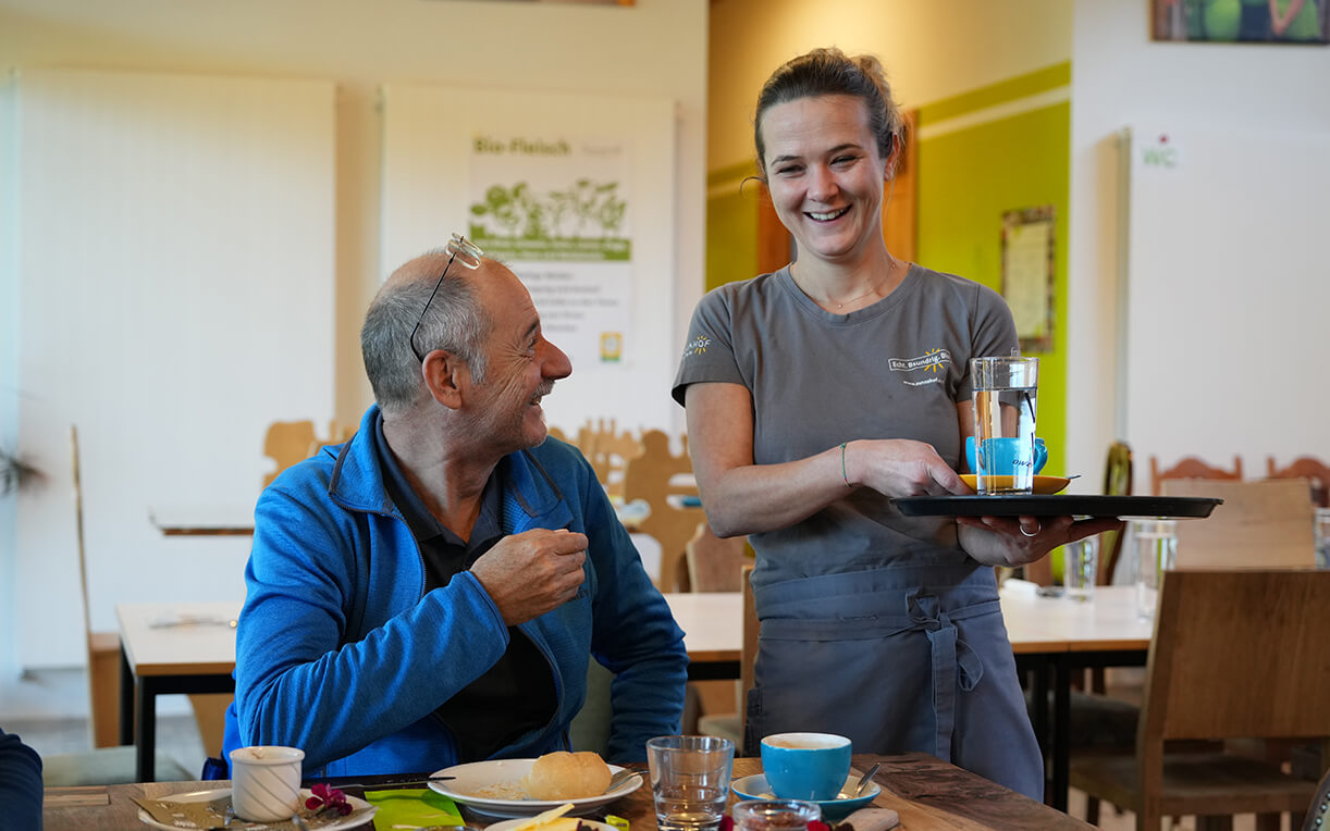 Eine fröhliche Kellnerin serviert einem lächelnden Mann am Tisch Getränke, während auf dem Tisch Speisen und Besteck liegen. Die Umgebung wirkt gemütlich und einladend, mit Holzmöbeln und sanfter Beleuchtung.