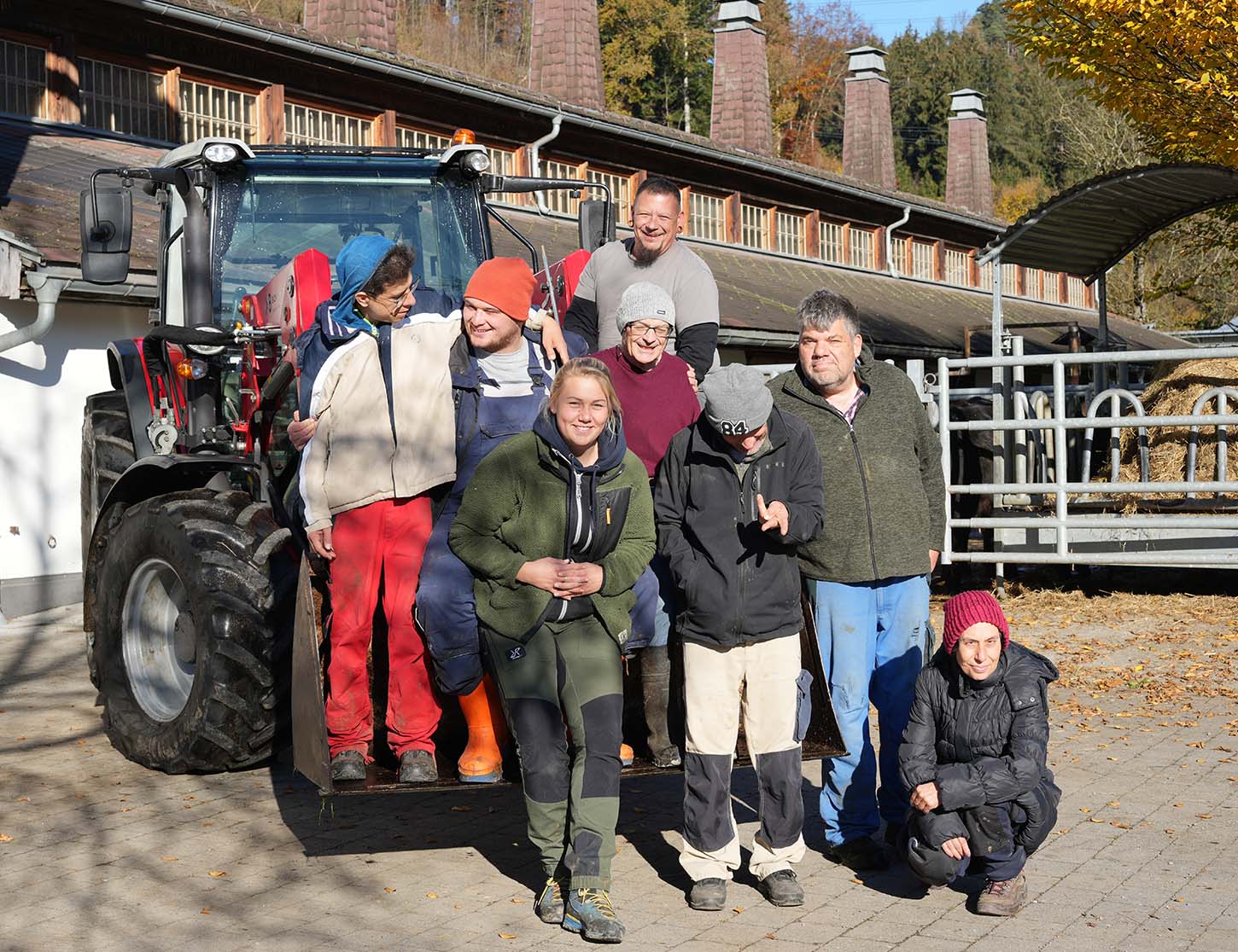 Eine Gruppe von acht Personen steht vor einem Traktor und einem Gebäude, lächelnd und in informeller Arbeitskleidung. Die Szene vermittelt ein Gefühl von Gemeinschaft und Freude. Im Hintergrund sind Bäume mit herbstlichen Farben zu sehen.