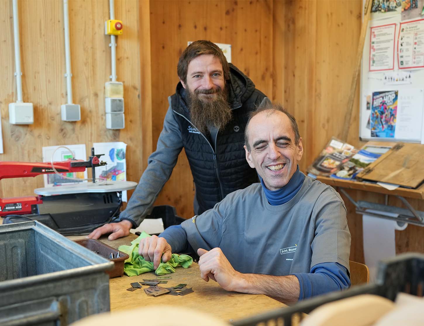 Zwei Männer stehen in einem Raum mit Holzverkleidung. Der Mann im Vordergrund sitzt und lächelt, während er auf einen Tisch zeigt, auf dem kleine Teile liegen. Der andere Mann steht hinter ihm und schaut freundlich in die Kamera.
