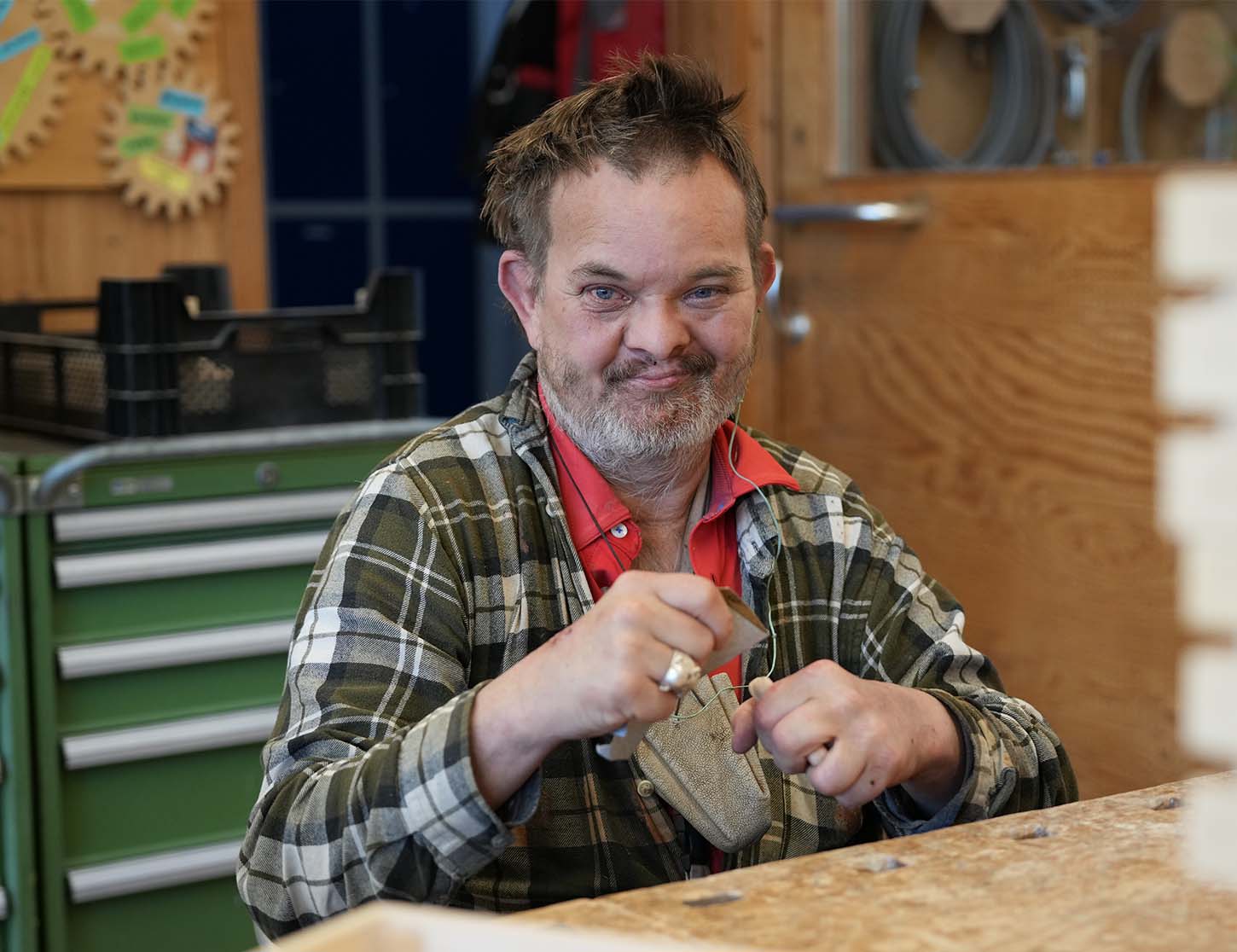 Ein Mann mit einem lächelnden Gesicht sitzt an einem Tisch und bearbeitet ein Holzstück mit einem Werkzeug. Er trägt ein kariertes Hemd und wirkt zufrieden mit seiner Arbeit. Im Hintergrund sind Werkzeuge und Materialien sichtbar, die auf eine Werkstatt hindeuten.