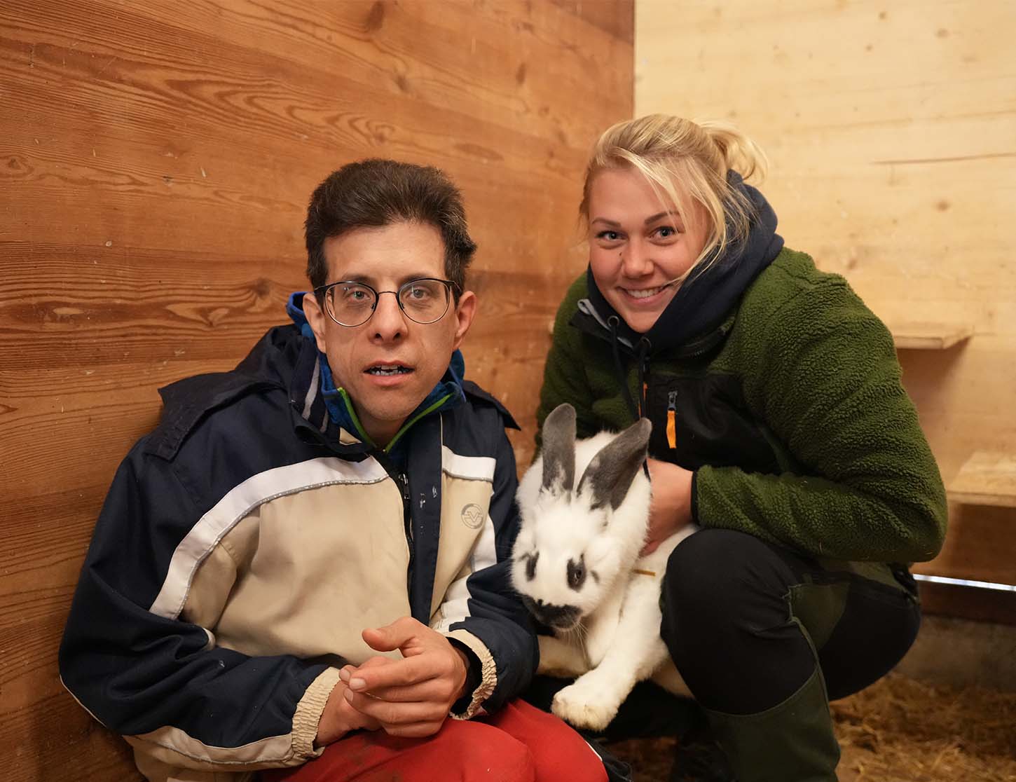 Eine Person sitzt an der Wand eines Stalls und hält die Hände in den Schoß. Neben ihr steht eine zweite Person, die ein Kaninchen im Arm hält. Beide lächeln in die Kamera. Der Hintergrund ist aus Holz und zeigt wenig weitere Details.