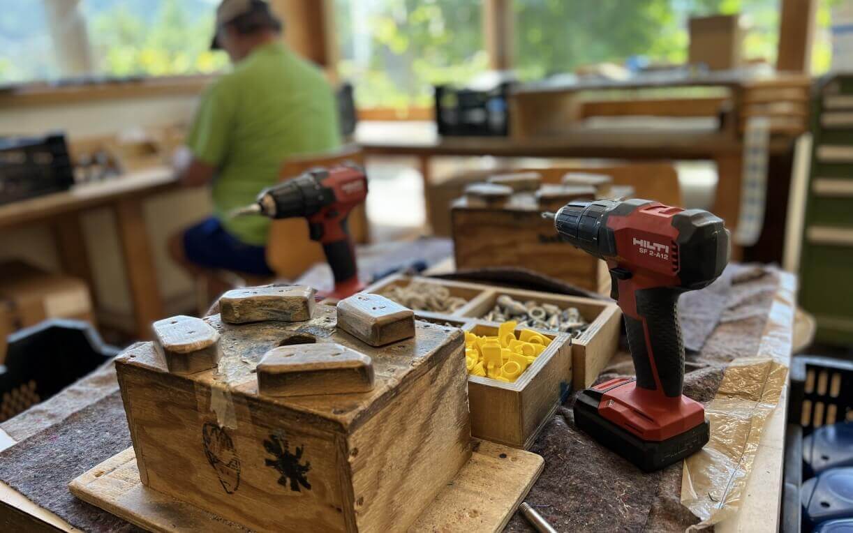 Auf einem Tisch im Vordergrund liegen zwei Akkuschrauber. Außerdem sind Holzboxen mit verschiedenen Bauteilen und Werkzeugen sichtbar, darunter Befestigungselemente in unterschiedlichen Farben. Im Hintergrund arbeitet eine Person an einem weiteren Tisch.