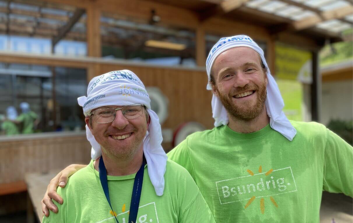 Zwei lächelnde Männer stehen nebeneinander, beide tragen grüne T-Shirts mit dem Schriftzug „Bsundrig“. Der Mann links hat eine Brille und trägt ein weißes Kopftuch, der andere hat einen Bart und ebenfalls ein weißes Kopftuch. Im Hintergrund sind unscharfe Personen und eine Holzkonstruktion sichtbar.
