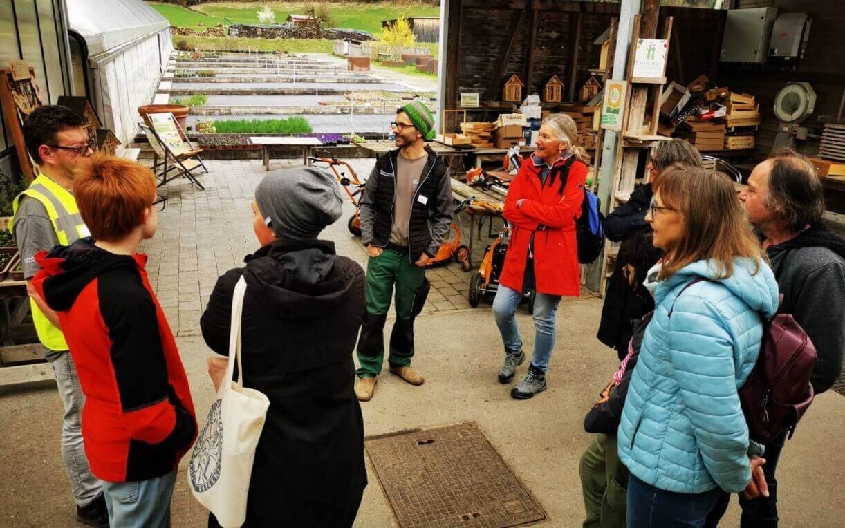Eine Gruppe von acht Personen steht auf einem Hof, während ein Mann mit Brille und Warnweste spricht. Die Teilnehmenden hören aufmerksam zu, einige lächeln. Im Hintergrund sind Tische mit Werkzeugen und Pflanzenbeeten zu sehen.