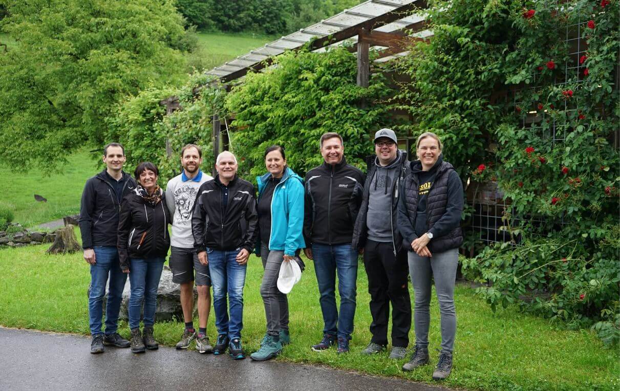 Eine Gruppe von acht Menschen steht auf einer Wiese vor einem grünen, mit Pflanzen bewachsenen Holzbau. Alle tragen Freizeitkleidung und lächeln. Die Szene vermittelt ein Gefühl von Freundschaft und Gemeinschaft in der Natur.
