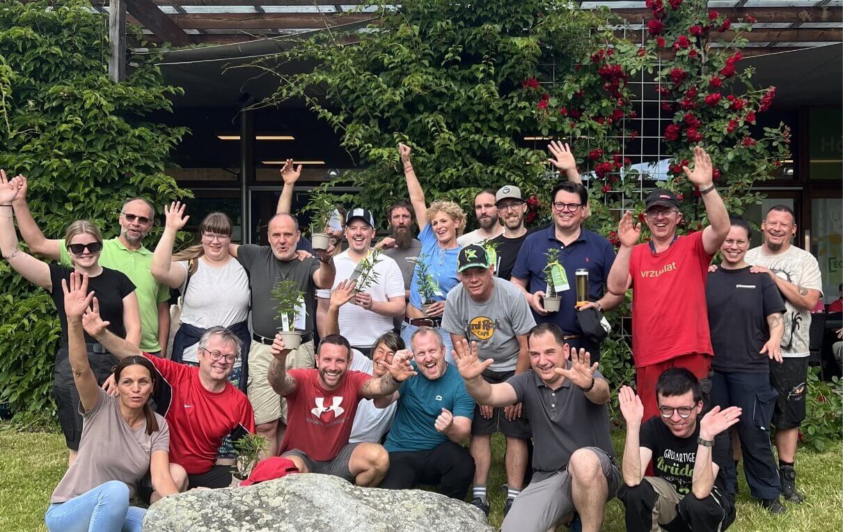 Eine Gruppe von etwa 20 Personen steht fröhlich zusammen. Viele halten kleine Pflanzen in Töpfen. Die Menschen winken lächelnd in die Kamera, umgeben von grünem Laub und blühenden Rosen. Die Atmosphäre ist freundlich und feierlich.