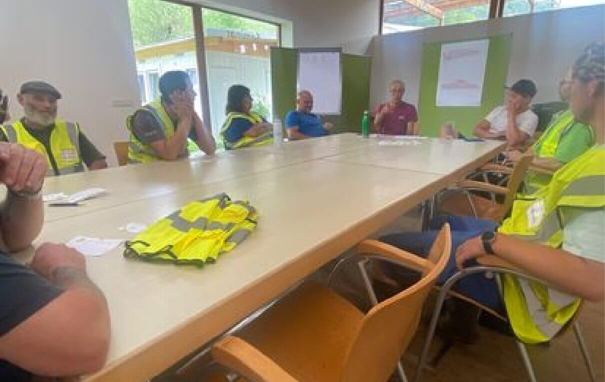 Eine Gruppe von Personen in leuchtenden Sicherheitswesten sitzt an einem Tisch in einem Besprechungsraum. Einige hören zu, während eine Person spricht. Auf dem Tisch liegen Unterlagen und eine gelbe Weste. Dahinter sind Fenster, die Tageslicht hereinlassen.