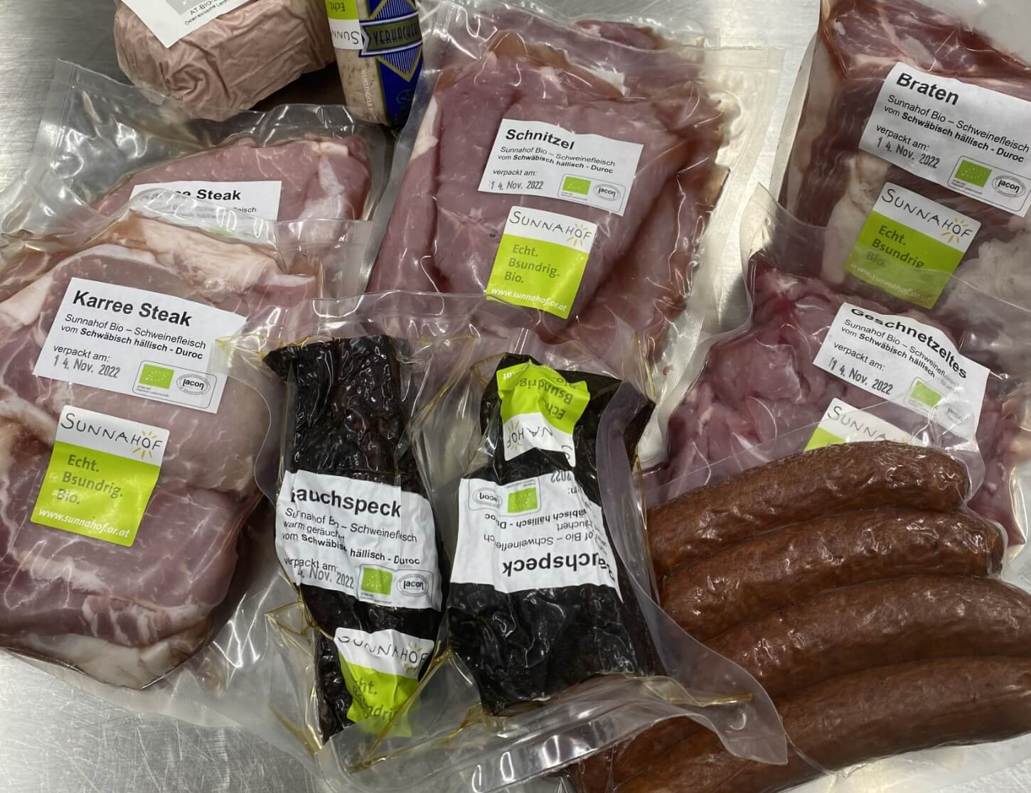 Auf dem Bild sind verschiedene verpackte Fleischprodukte zu sehen, darunter Karree Steak, Schnitzel, Braten, Speck und Würste. Alle Verpackungen stammen von einem Bio-Lieferanten und enthalten Beschriftungen mit den Produktnamen und dem Verpackungsdatum.